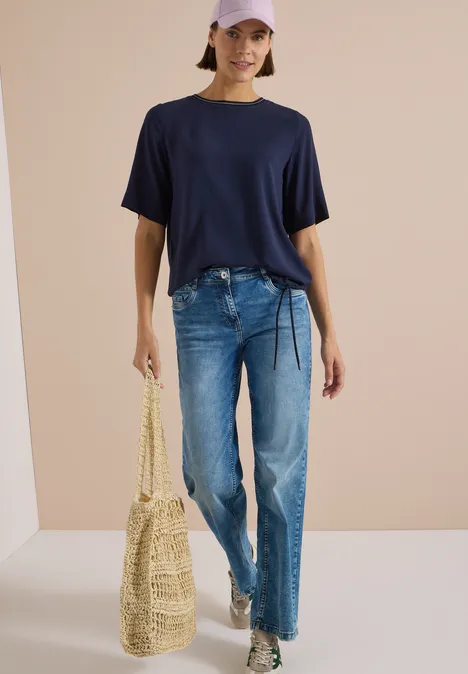 Blouse met korte mouwen en ronde hals urban dark blue Frau in lässigem Outfit: dunkelblaues T-Shirt, Jeans, weiße Sneaker, Strohtasche und rosa Cap.