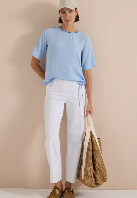 Frau in hellblauem T-Shirt, weißer Hose, Strohtasche und Cap vor beigem Hintergrund.