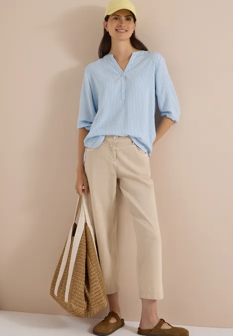 Frau in lässigem Sommeroutfit: hellblaue Bluse, beige weite Hose, Sandalen, Strohtasche und gelbe Kappe.