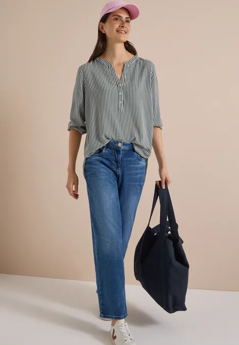 Frau in grauem Oberteil, blauen Jeans, rosa Mütze und schwarzer Tasche vor beigem Hintergrund.