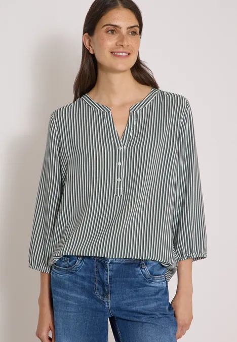 Frau mit grüner, gestreifter Bluse und blauen Jeans vor hellem Hintergrund.