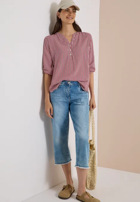 Frau lehnt lässig an Wand, trägt lila Bluse, weite Jeans, Cap, Sandalen und Basttasche.