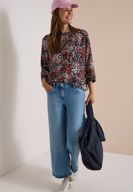 Blouse met 3/4-mouwen en print raw chocolate brown Vrouw lacht in gebloesd shirt, wijde spijkerbroek, roze pet, witte sneakers, draagt een donkerblauwe tas.