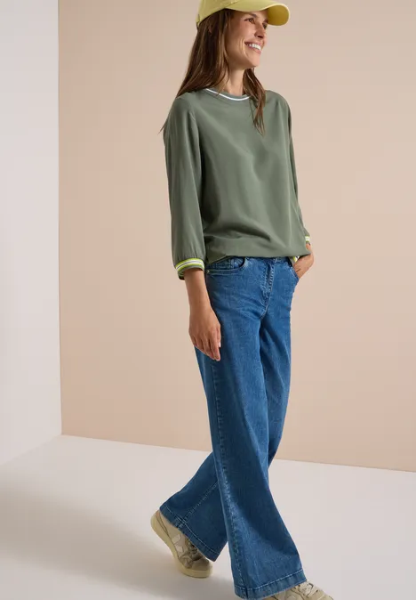 Blouse met 3/4-mouwen en contrasterende details dried olive green Vrouw met gele pet, groen truitje, wijde jeans en doorzichtige sneakers voor een neutrale achtergrond.