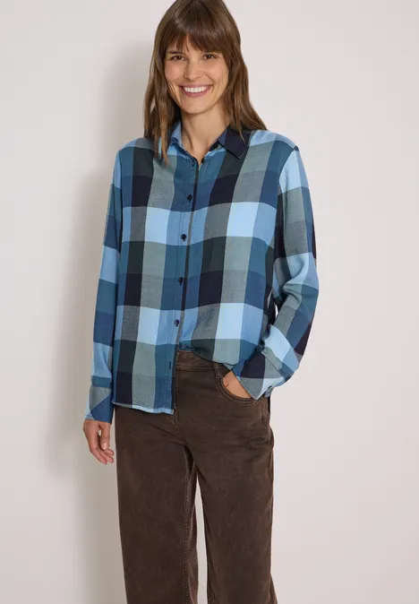 Eine lächelnde Frau trägt ein blau kariertes Hemd und braune Hose vor hellem Hintergrund.