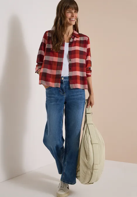 Junge Frau in karierter roter Hemdjacke, weißem Top, blauen Jeans und großer beiger Tasche lächelt.