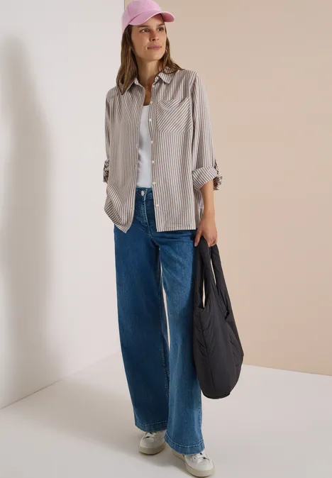 Frau in hellem Overshirt, weißem Shirt und weiten Jeans, mit rosa Cap und schwarzem Beutel auf neutralem Hintergrund.