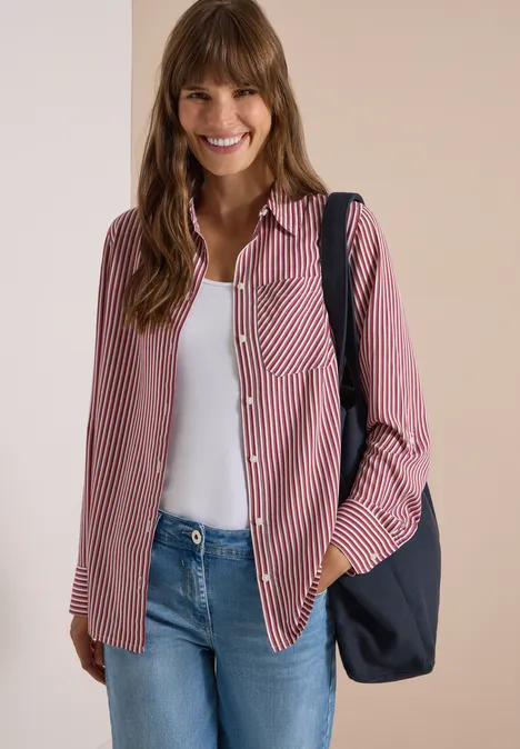 Fröhliche Frau trägt gestreiftes rotes Hemd über weißem Top, Jeans und schwarzen Schulterbeutel.