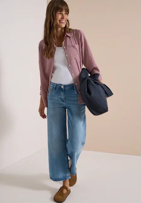 Junge Frau in gestreiftem Hemd, weißem Top, weiten Jeans und braunen Slippern, mit dunkelblauer Tasche.