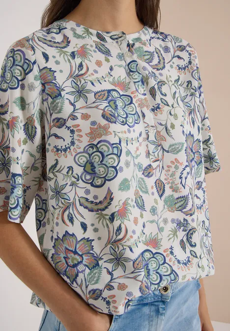 Frau mit weißer Bluse im blau-grünen Blumenmuster und Jeans vor hellem Hintergrund.