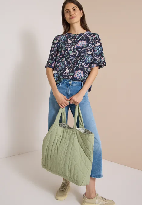Frau in gemusterter Bluse, Jeans und mit großer gesteppter grüner Tasche.