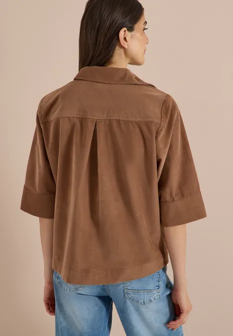 Overhemd met korte mouwen in effen kleur creamy mocha Vrouw van achteren in een losse bruine corduroy blouse met driekwartmouwen en blauwe spijkerbroek voor een beige achtergrond.