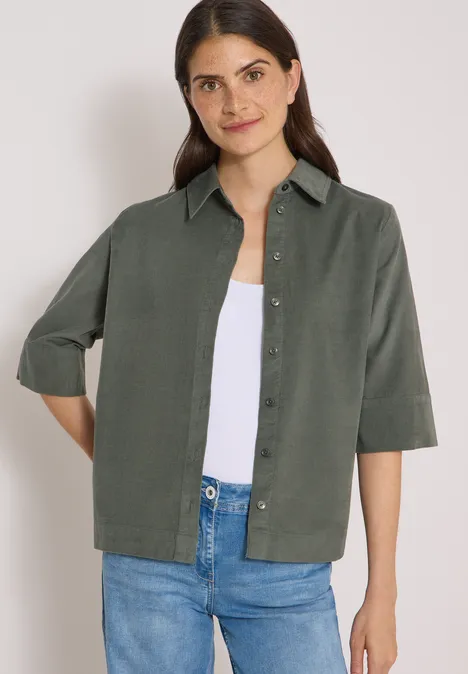 Overhemd met korte mouwen in effen kleur dried olive green Vrouw in groen overhemd met driekwartmouwen, wit topje en blauwe spijkerbroek, lichte glimlach.