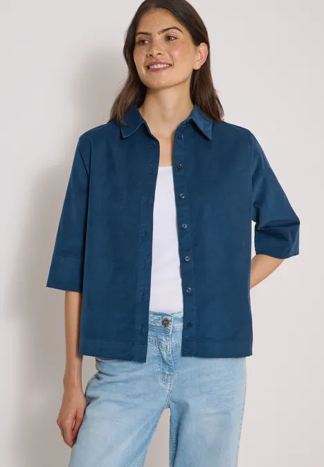 Frau trägt blaues Jackett über weißem Top und Jeans, steht vor neutralem Hintergrund.