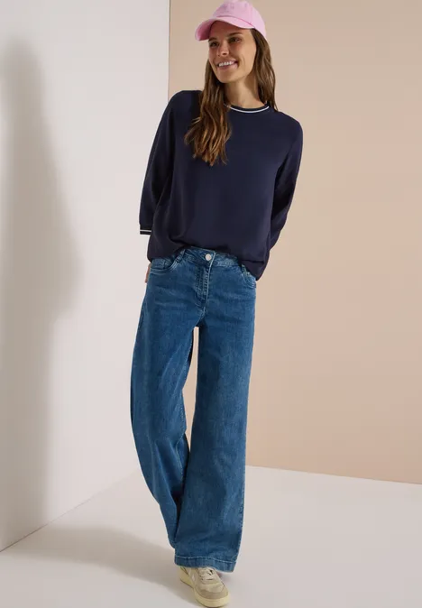 Frau in weitem Jeans, marineblauem Pullover und rosa Cap, lächelnd vor hellem Hintergrund.
