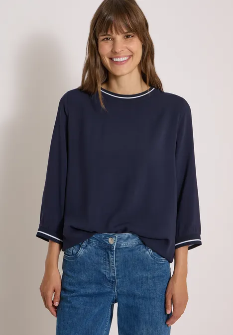 Eine lächelnde Frau trägt eine dunkelblaue, weite Bluse und Jeans.