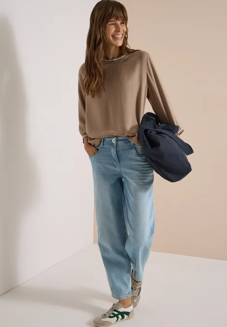 Eine Frau trägt eine braune Bluse, helle Jeans und grüne Sneaker, hält eine Jacke in der Hand.