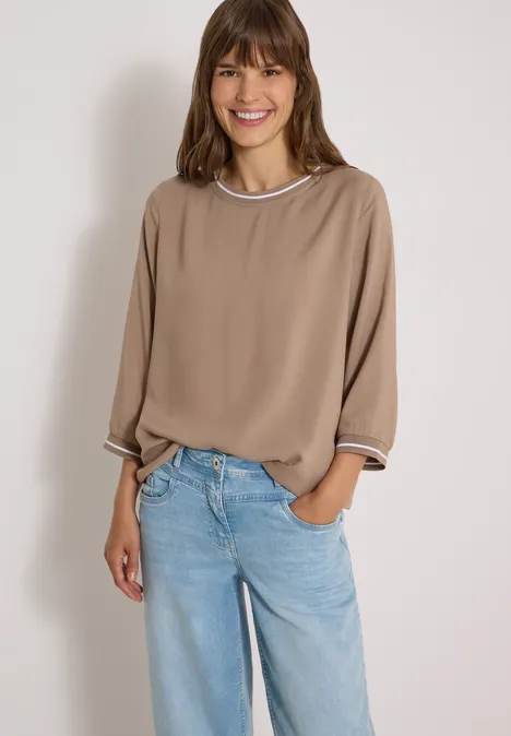Eine lächelnde Frau trägt eine beige Bluse und hellblaue Jeans vor weißem Hintergrund.