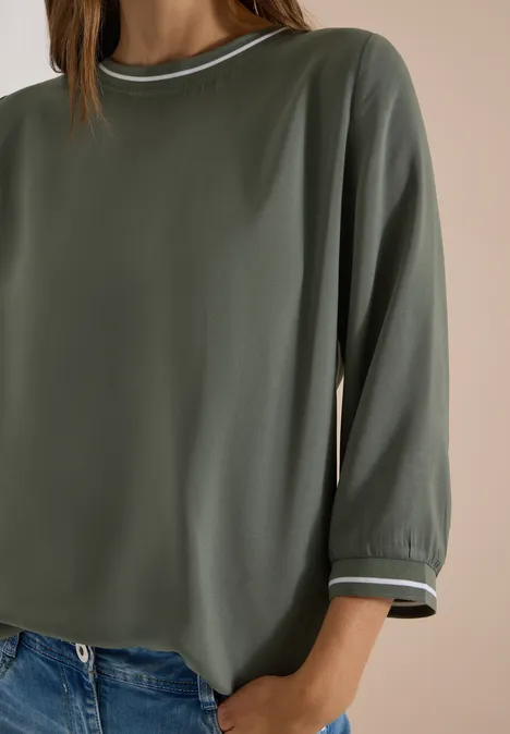 Eine lockere, olivgrüne Bluse mit 3/4-Ärmeln und weißem Paspel am Kragen und Saum.