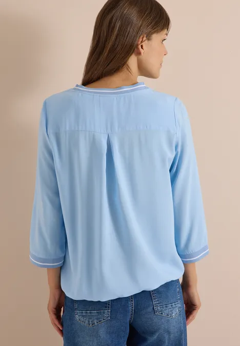 Rückansicht einer Frau in hellblauer Bluse mit 3/4-Ärmeln und Jeans, vor beigem Hintergrund.