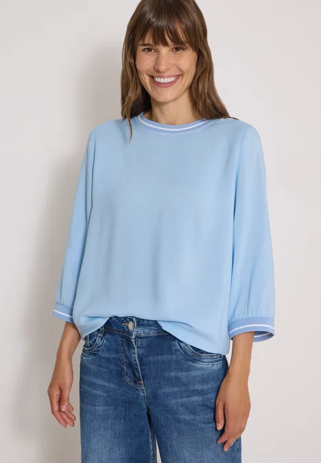 Lächelnde Frau in blauer Bluse und Jeans vor hellem Hintergrund.