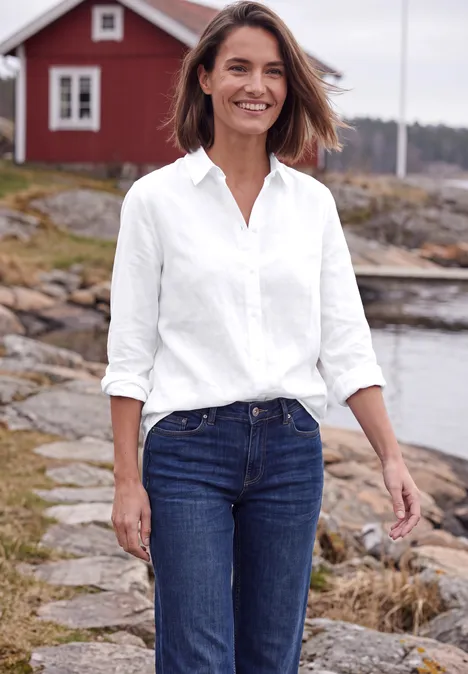 Langarm Hemdbluse mit Knopfleiste White Lächelnde Frau in weißem Hemd und Jeans spaziert auf felsigem Pfad am Wasser, rotes Haus im Hintergrund.