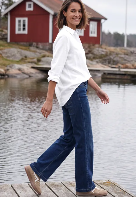 Langarm Hemdbluse mit Knopfleiste White Eine Frau in weißem Hemd und Jeans geht lächelnd auf einem Steg vor einer roten Hütte am Wasser.