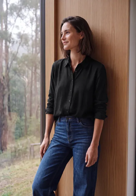Een glimlachende vrouw in een zwart overhemd en jeans leunt tegen een houten wand en kijkt naar het groen.
