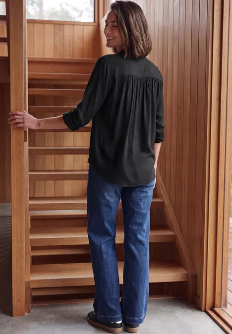 Vrouw in donkere blouse en spijkerbroek loopt een houten trap op in een warme, houten omgeving.