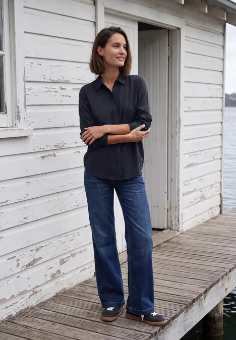 Langarm Hemdbluse mit Knopfleiste urban dark blue Eine Frau steht lächelnd in Bluse und Jeans vor einem weißen Haus am Wasser.