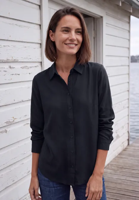 Langarm Hemdbluse mit Knopfleiste urban dark blue Eine lächelnde Frau in schwarzer Bluse und Jeans steht auf einem Steg vor einem weißen Holzhaus am Wasser.