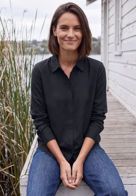 Langarm Hemdbluse mit Knopfleiste urban dark blue Eine Frau sitzt auf einem Holzsteg am Wasser, umgeben von Schilf, in Jeans und schwarzer Bluse lächelnd.