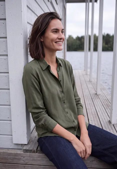 Een glimlachende vrouw in een groen overhemd zit ontspannen op een houten steiger aan het water.