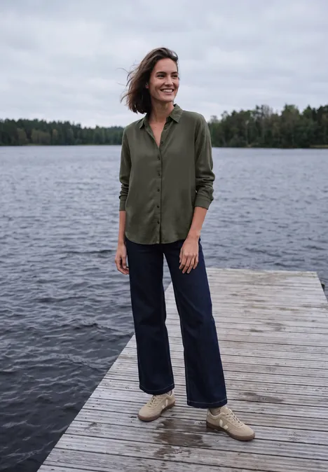 Een vrouw staat op een houten steiger aan het meer, gekleed in een groen overhemd, jeans en lichte schoenen.