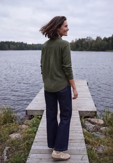 Vrouw in groen overhemd en donkere broek glimlachend op een steiger bij een meer, omgeven door bomen.