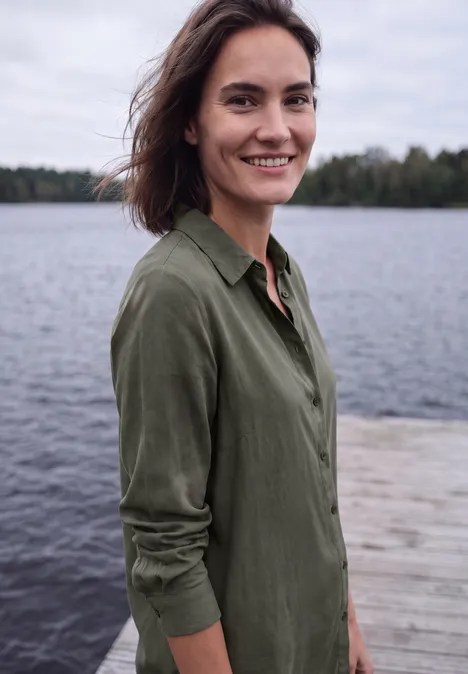 Een glimlachende vrouw staat op een steiger aan een meer, draagt een groen overhemd en kijkt in de camera.
