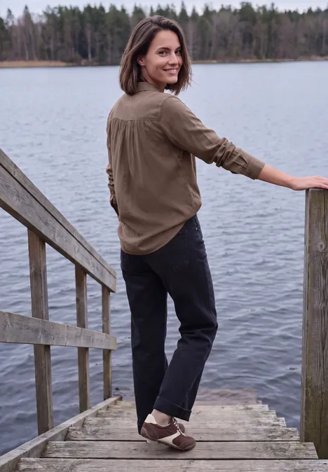 Langarm Hemdbluse mit Knopfleiste urban taupe Eine Frau steht lächelnd auf einem Steg an einem See, trägt braunes Hemd und schwarze Hose.