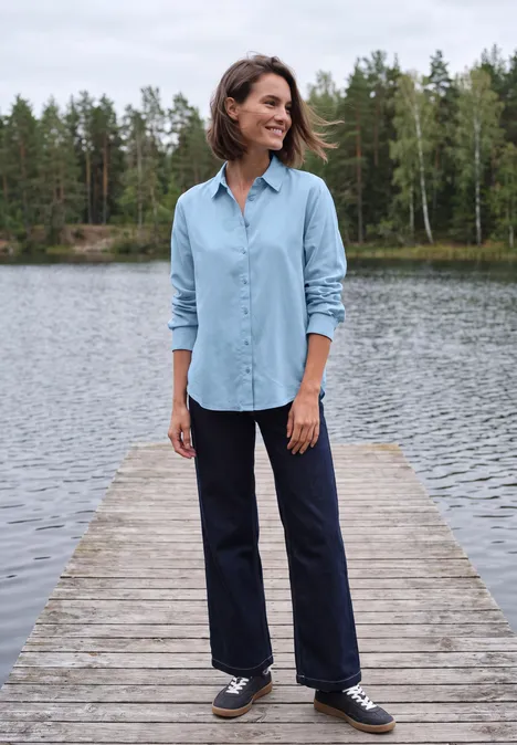 Vrouw staat glimlachend op een steiger bij een meer, draagt lichtblauw shirt en donker broek, bomen op de achtergrond.