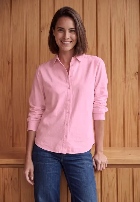 Langarm Hemdbluse mit Knopfleiste mellow rose Frau mit rosa Hemd und Jeans steht lächelnd vor einer Holzwand.