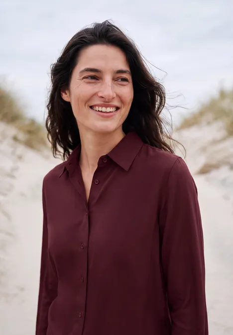 Chemisier à manches longues avec patte de boutonnage red wine Eine Frau lächelt vor Dünen am Strand.