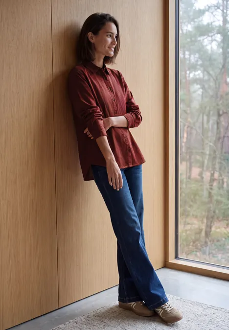 Langarm Hemdbluse mit Knopfleiste russet red Eine lächelnde Frau lehnt in einem roten Hemd an einer Holzwand und blickt aus dem Fenster in den Wald.