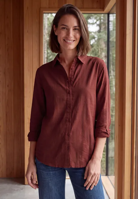 Langarm Hemdbluse mit Knopfleiste russet red Eine lächelnde Frau in rotem Hemd und Jeans steht in einem modernen Holzhaus vor großen Fenstern.