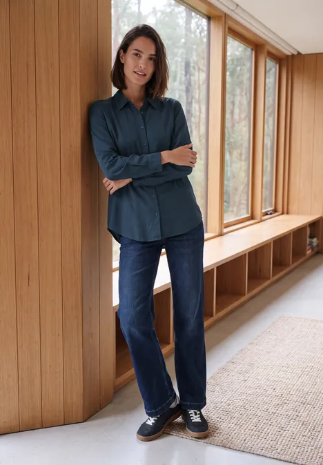Langarm Hemdbluse mit Knopfleiste moonlight blue Frau in Jeans und blauem Hemd lehnt lässig an einer Holzwand vor großen Fenstern.