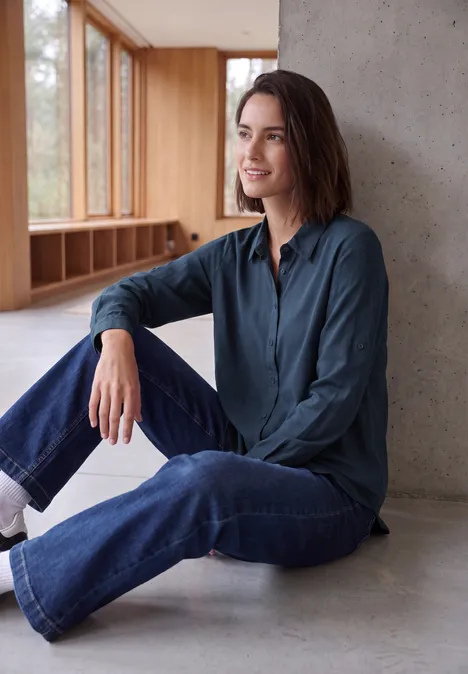 Langarm Hemdbluse mit Knopfleiste moonlight blue Junge Frau sitzt lässig im modernen Raum, lehnt an einer Betonwand, trägt Jeans und dunkelblaue Bluse.
