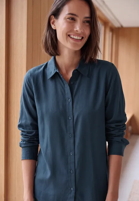 Langarm Hemdbluse mit Knopfleiste moonlight blue Lächelnde Frau mit dunkelblau-grüner Bluse in hellem Raum vor Holzwänden.