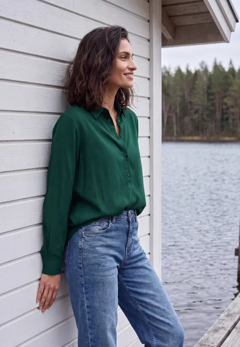 Een vrouw in een groene blouse en spijkerbroek leunt tegen een witte houten wand met een meer en bos op de achtergrond.