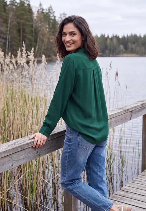 Vrouw in groen overhemd lacht op houten steiger aan een meer, omringd door riet en bos op de achtergrond.