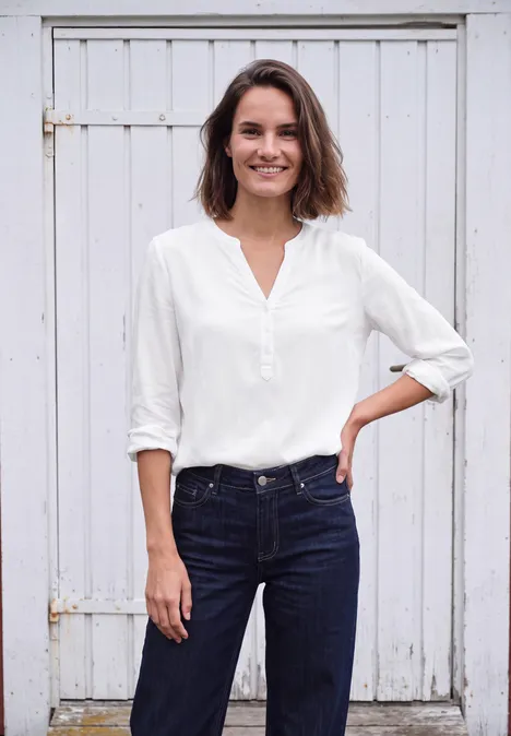 Langarm Tunika mit Split Neck White Frau in weißer Bluse und Jeans vor weißem Holztor, lächelt und hat die Hand in der Hüfte.