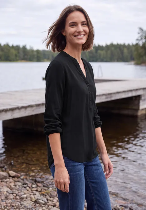 Een vrouw in zwarte blouse en spijkerbroek glimlacht op een steiger bij een rustig meer.