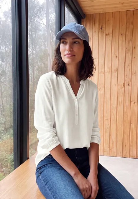 Frau mit weißer Bluse, Jeans und Cap sitzt entspannt an einem Fenster in einem Holzinterieur.